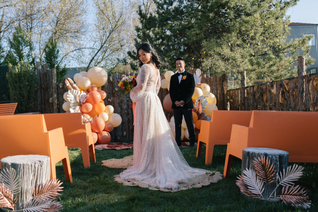 Campfire Hotel orange plastic benches at the ceremony site for a modern bride and groom. Orange and white balloons behind the groom who waits for his bride who is mid-aisle.