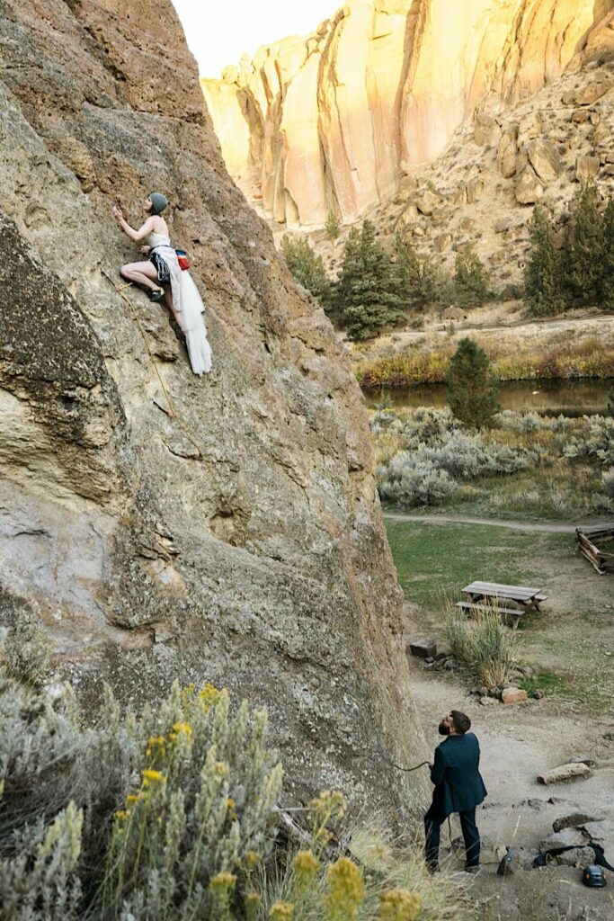 Bride ascends rock face and groom maintains rope. 