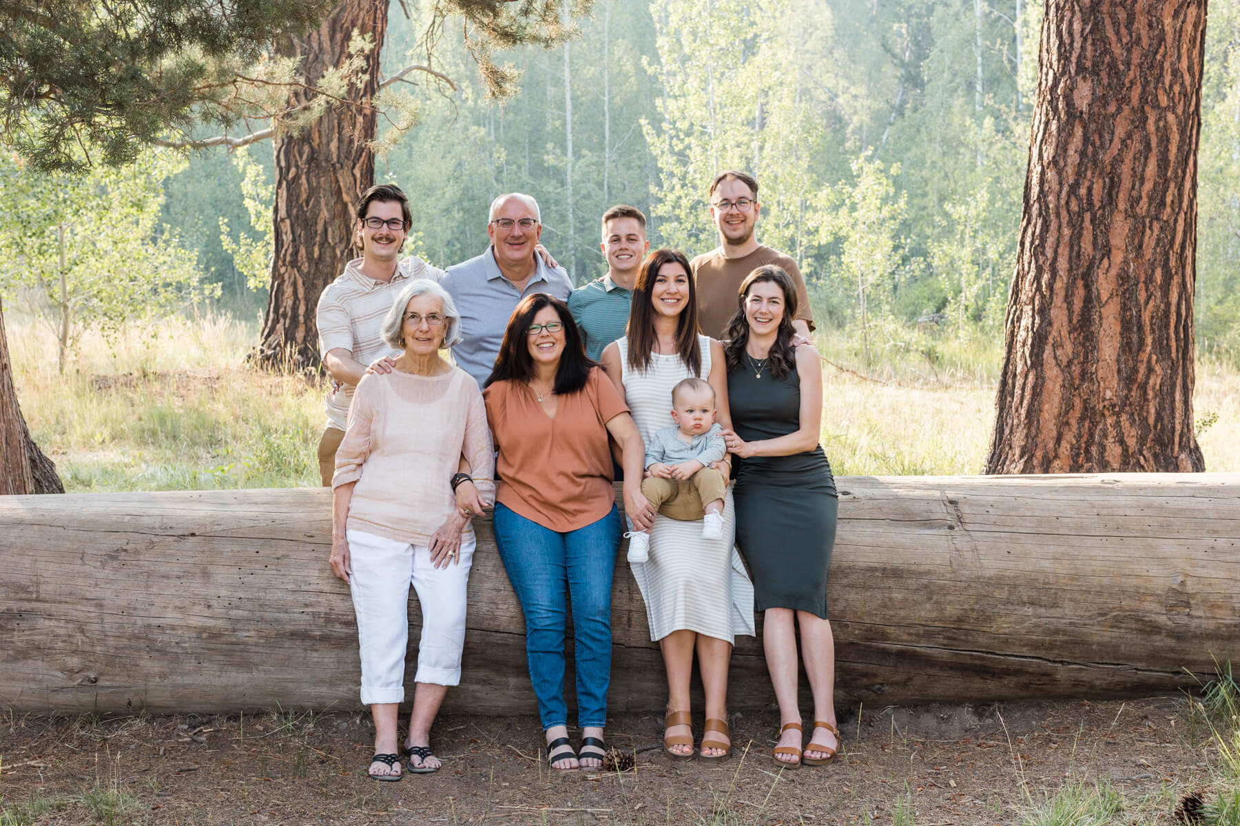 Family photo of four generations sitting on a large downed log in a wooded park. 
