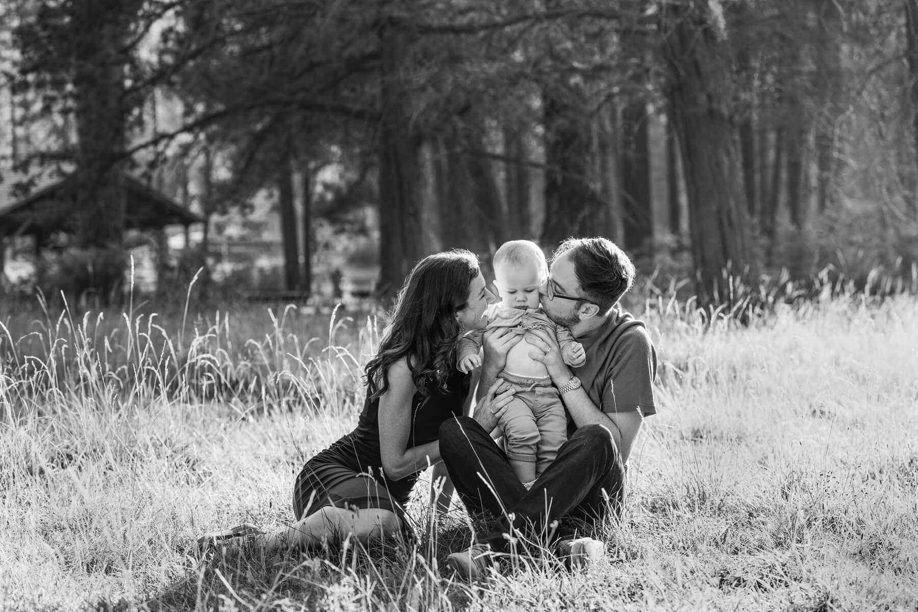 Black and white image of new parents sitting in tall grass kissing their baby. 