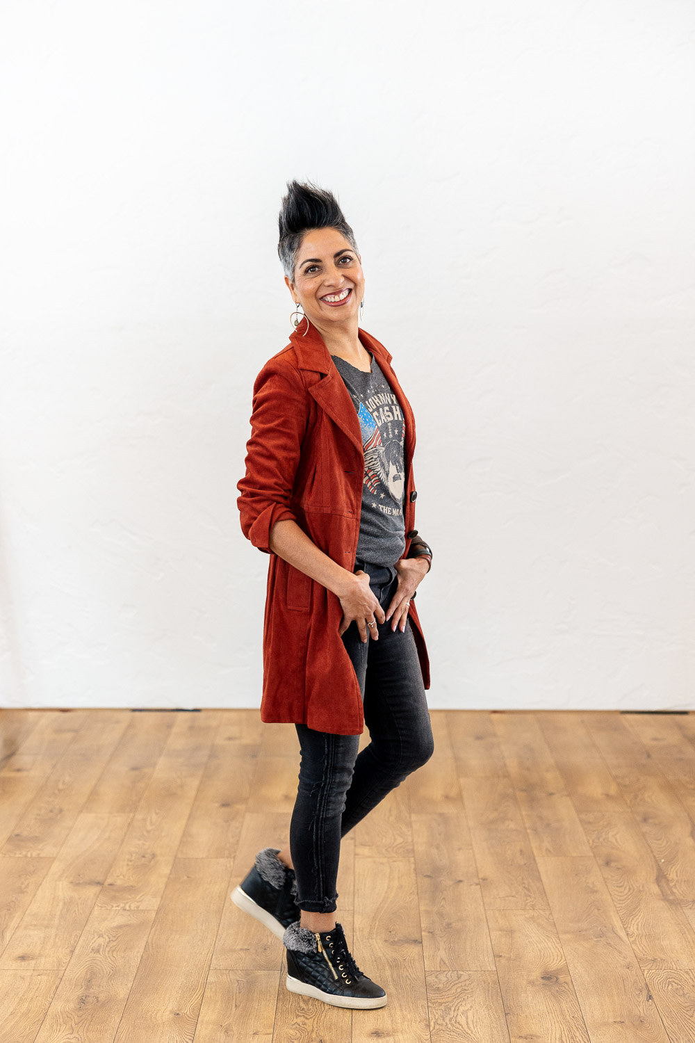 Woman with short dark styled hair smiles at camera, wearing rust colored coat, dark jeans and sneakers.