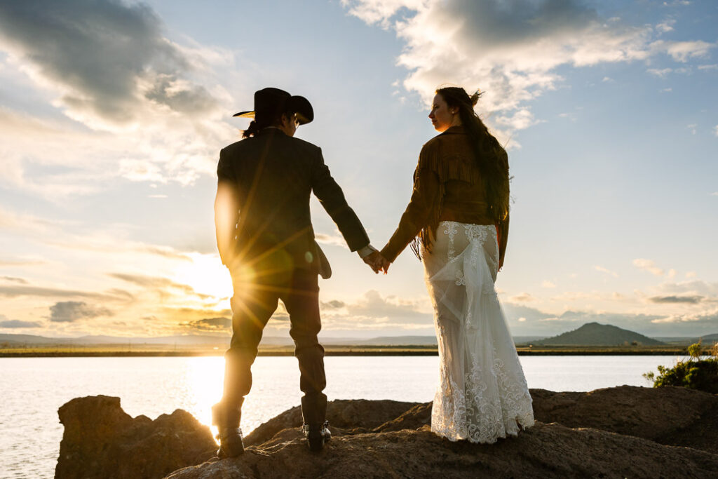 Couple standing on rocks holding hands at sunset. water and clouds in the background.
