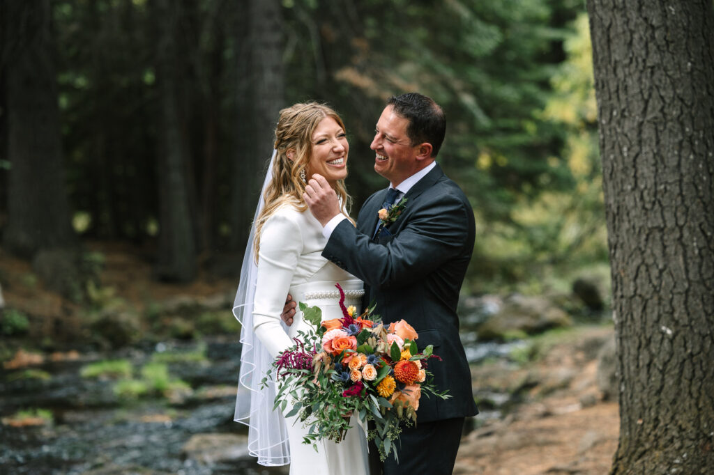 A bride has a large smile on her face as her smiling groom brushes a strand of hair off her face. The couple stands by a stream in the woods.