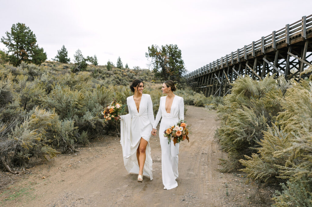 Two brides walk hand in hand on dirt path.