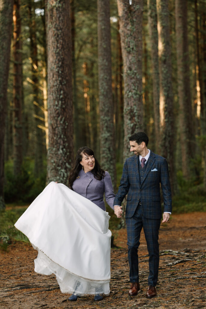 Newly weds walk hand in hand smiling at each other through heavily wooded trees in the Cairngorms in Scotland.