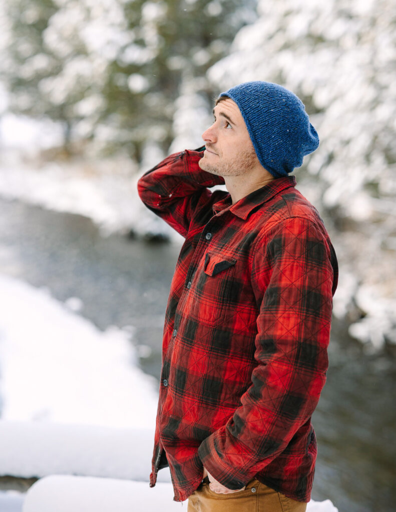 Man in red plaid shirt and blue beanie made by Wool Town. Background is blurred snowy trees and winter stream.