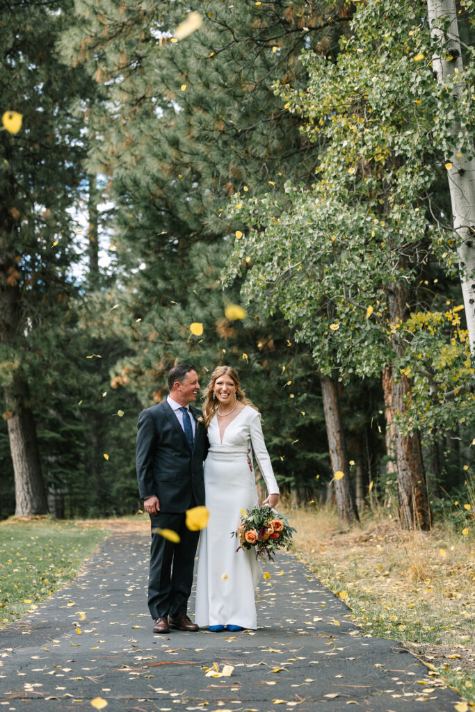 Bride and groom stops on bike path as a gust of wind blows golden aspen leaves around them. 