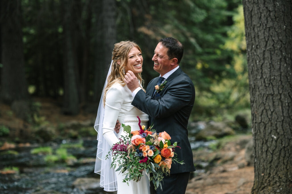 Candid moment of the smiling couple near a creek. Bride is holding brightly colored bouquet with oranges, coral and maroon colors. 
