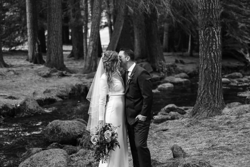 Black and white image of groom kissing bride's cheek in forest.