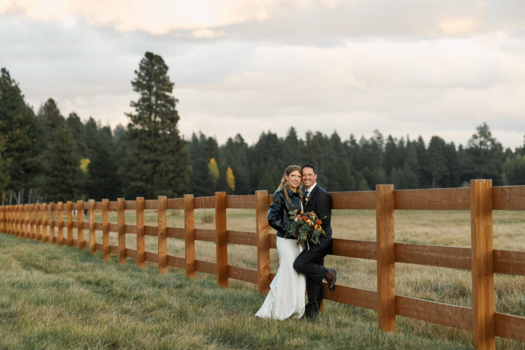 Newlyweds lean in together against a wooden fence in a field whilte smiling at the camera. 