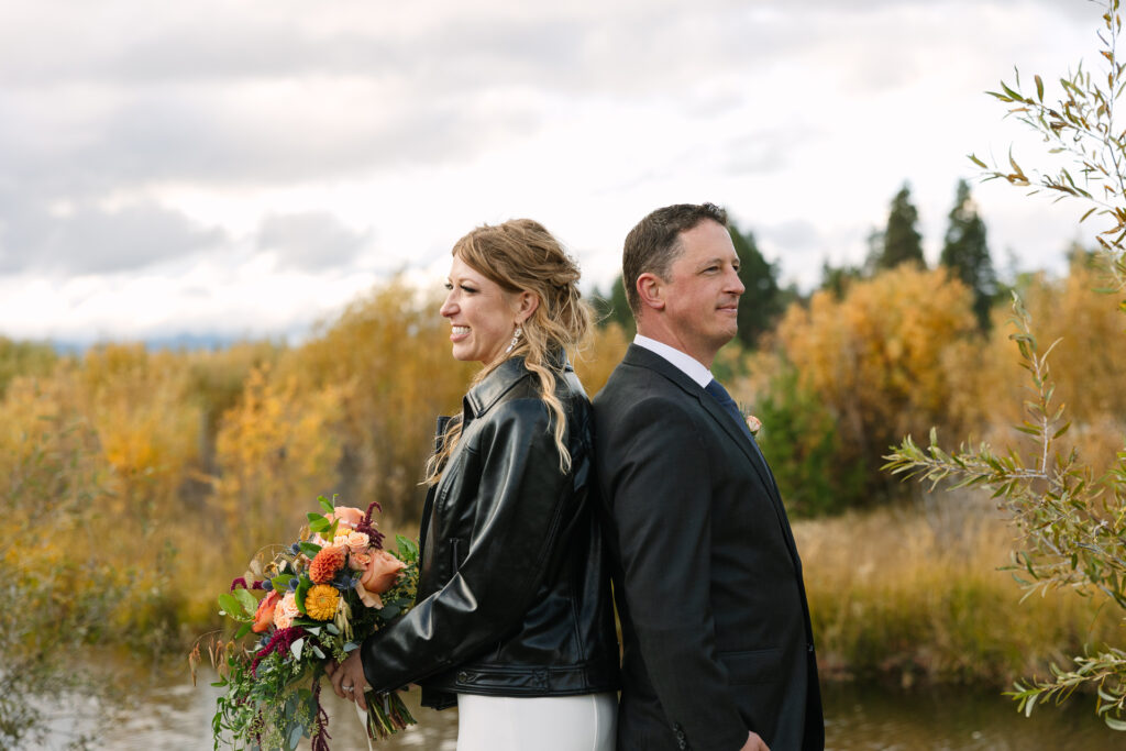 Bride wears black leather jacket, holding her bouquet standing back to back with her husband near the creek and golden shrubs. 