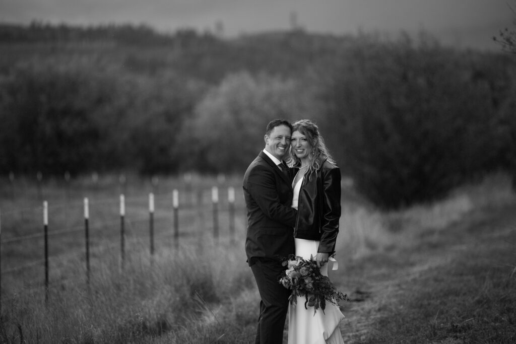 Moody black and white portrait of the couple leaning in smiling. Bride holds bouquet down by waist, wearing leather jacket. 