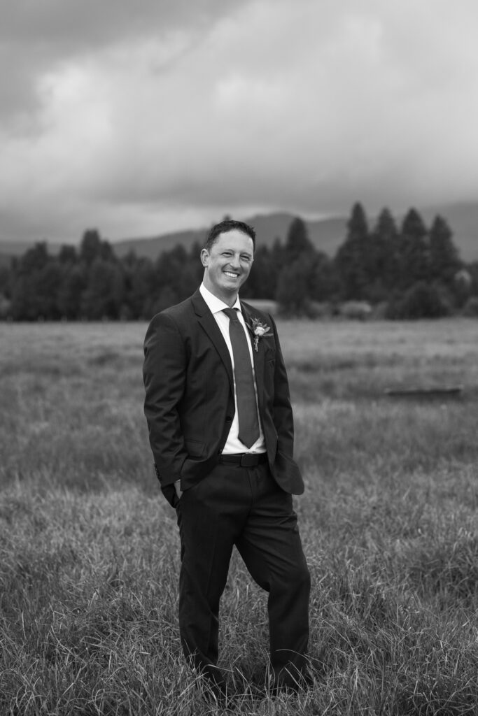 Full length moody black and white portrait of groom smiling in field. 