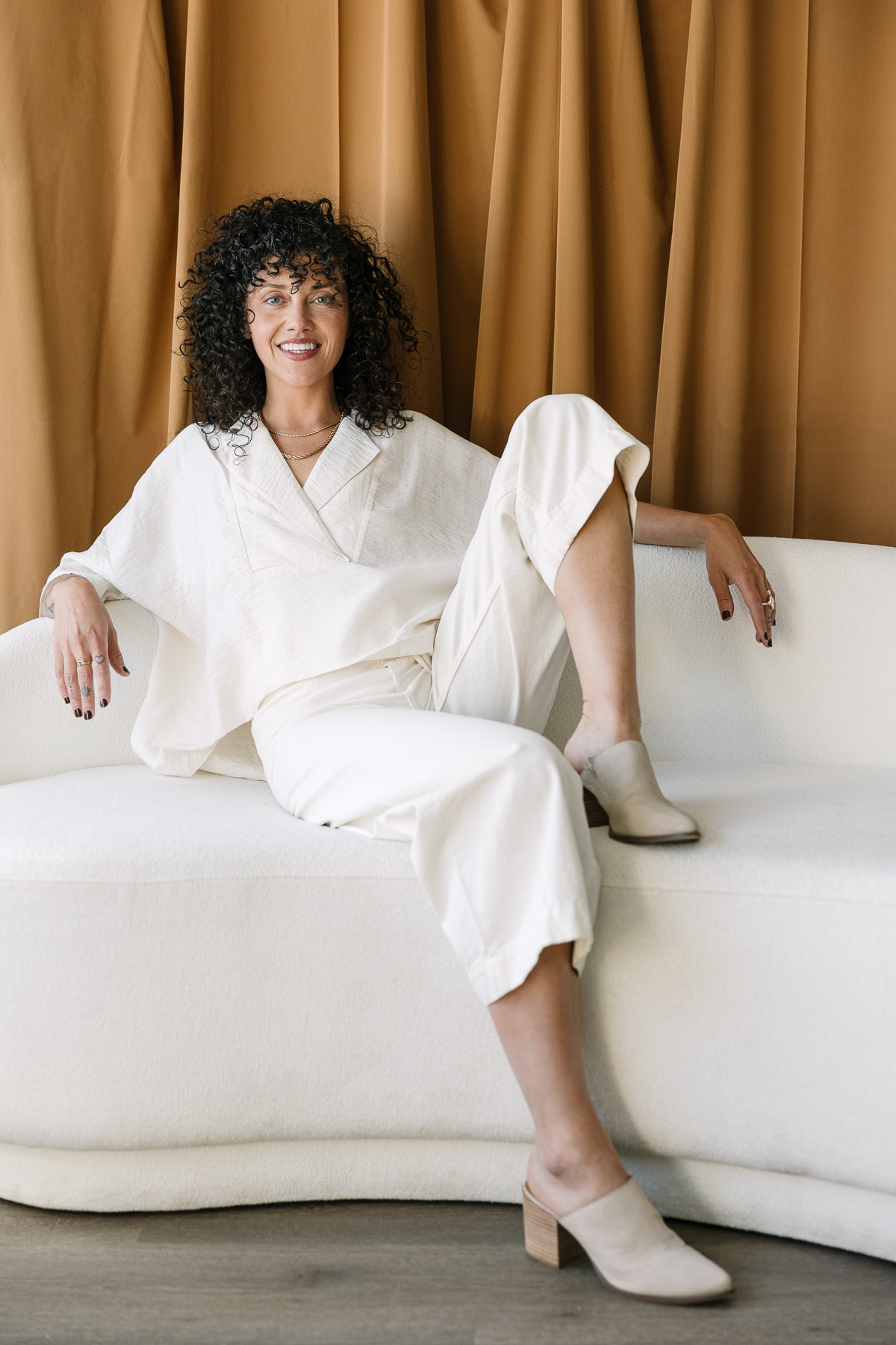 Woman in all white sits on white couch with brown fabric behind her.