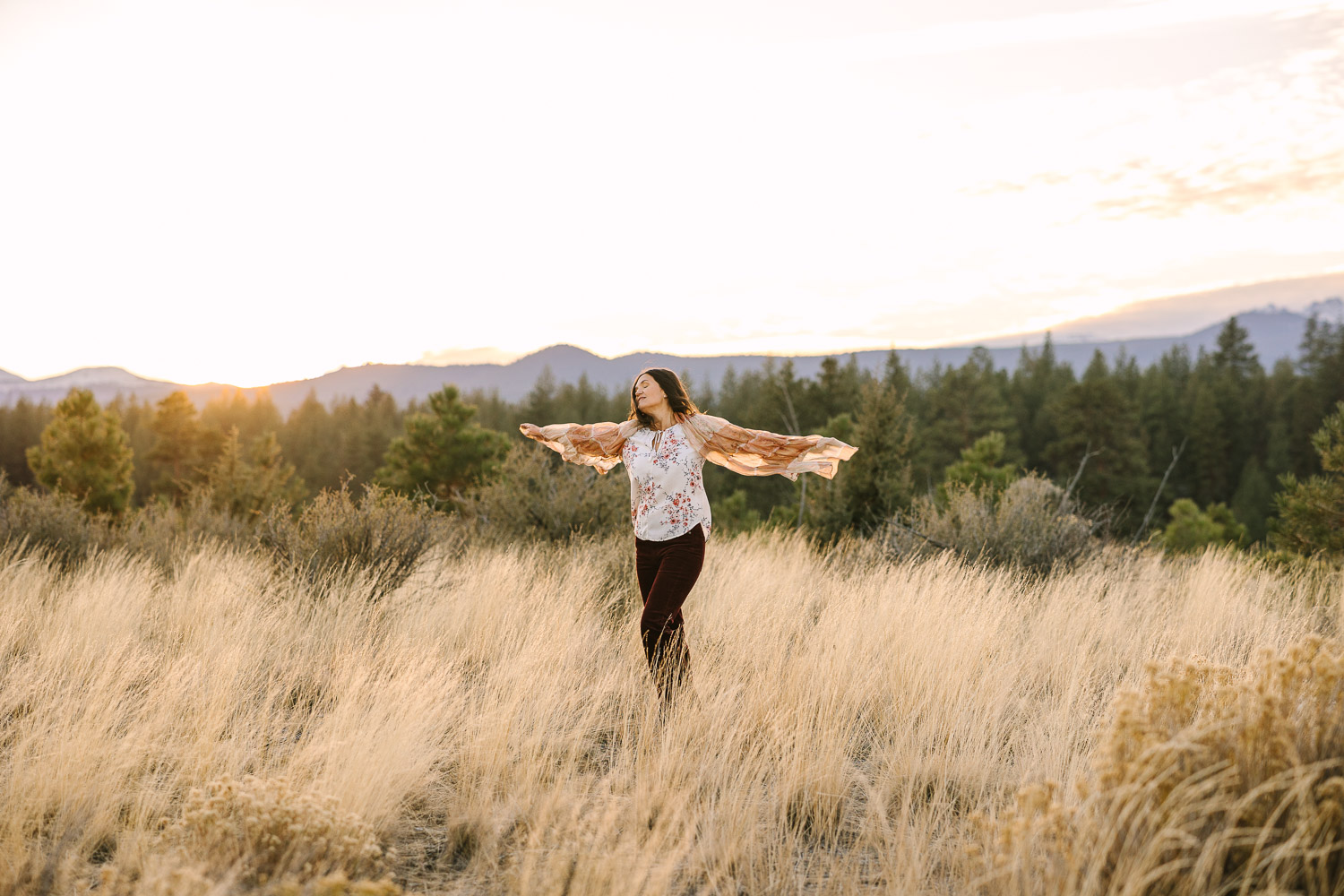 Brunette woman stands in golden field with outstretch arms holding scarf.