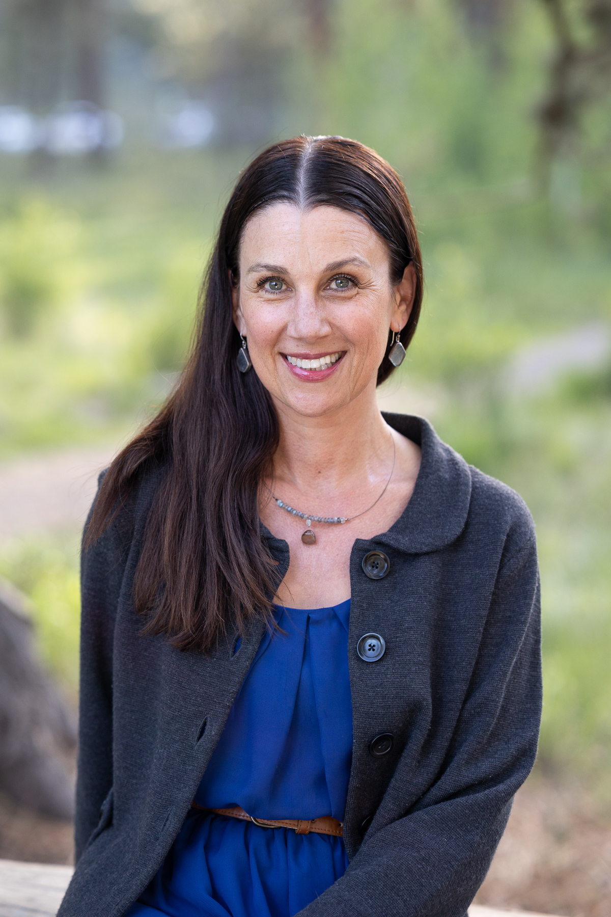 Woman with dark hair blue dress and grey sweater smiles at the camera. 