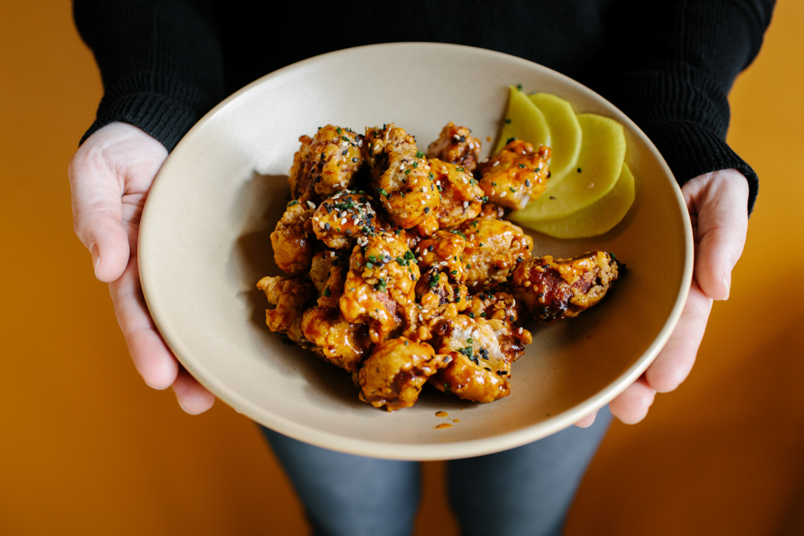 Gluten free korean chicken at Mill Works Pub. Chicken is in a bowl being held against an orange wall that matches the chicken's color.