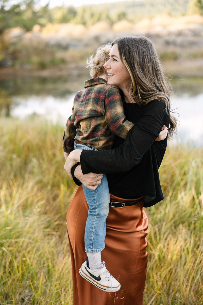 Mother in black sweater and rust colored skirt holds young toddler near a river.