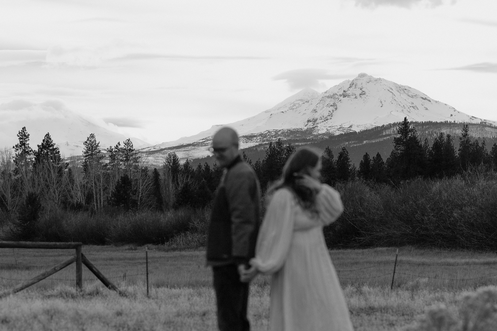 couple holds hands with snow covered mountains in the distance. Image is black and white.
