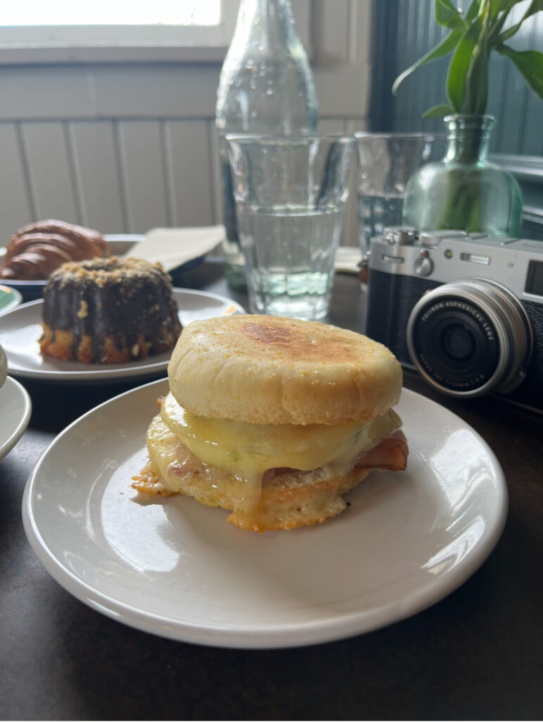 Gluten free english muffin egg sandwich on table with camera and mini bundt cake in the background.