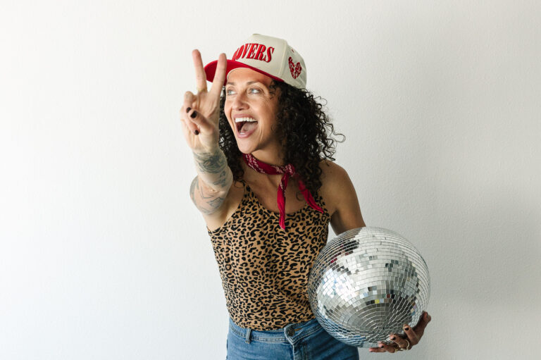 Women wearing ball cap, holding disco ball gives peace sign.