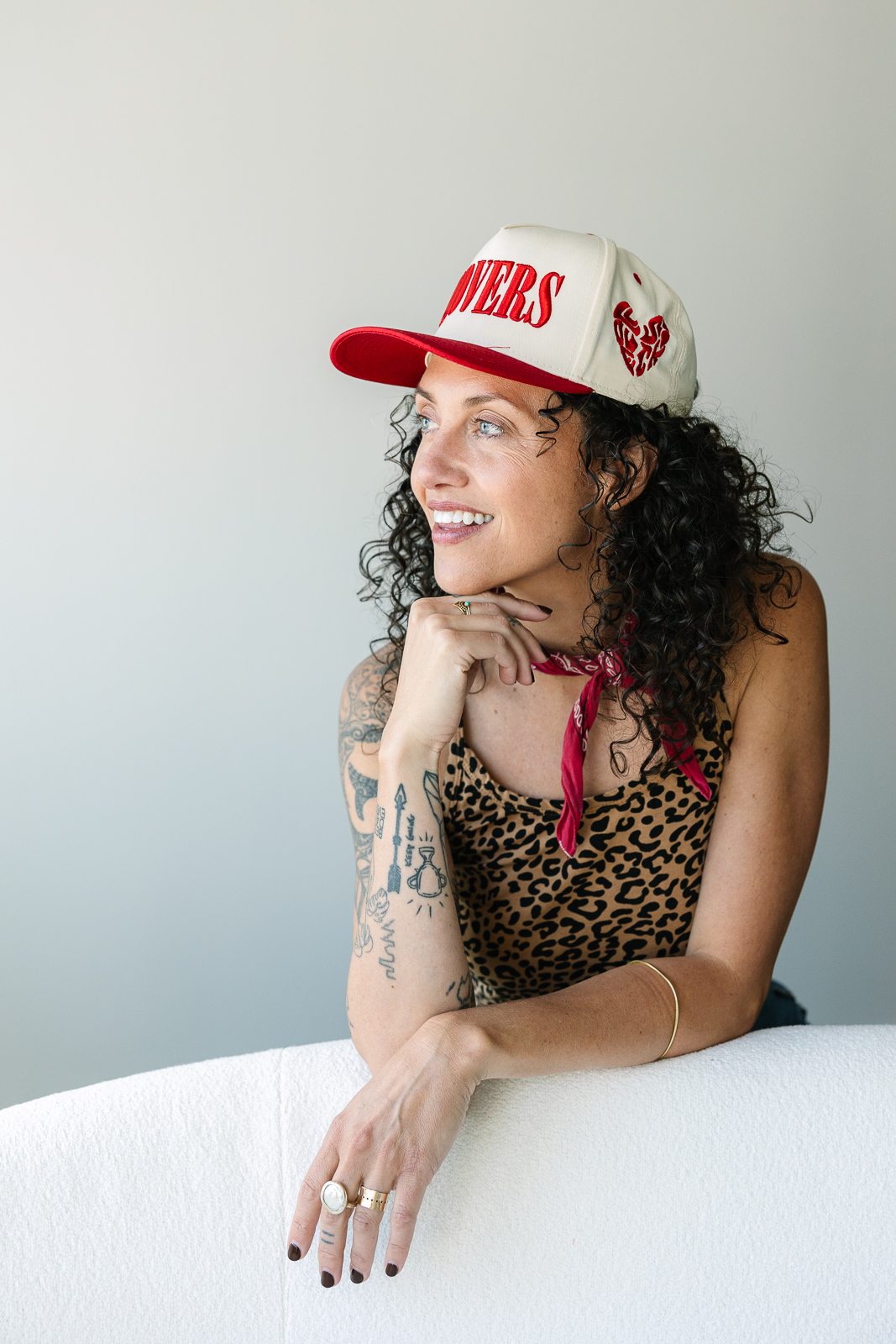 Brunette wearing a cream and read ball cap leans on white couch and smiles.
