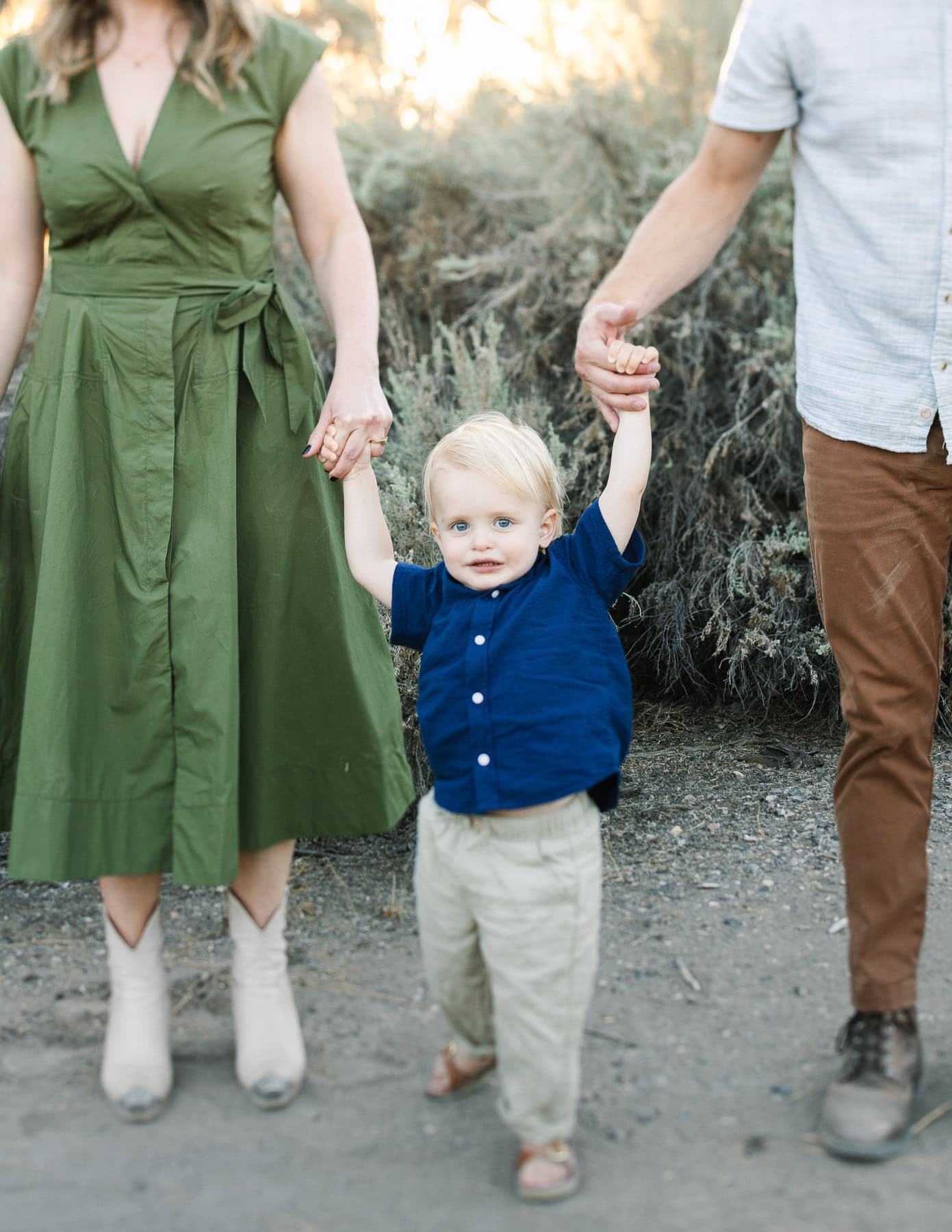 closeup image of a toddle boy in a blue shirt and tan pants holding mom and dad's hands.