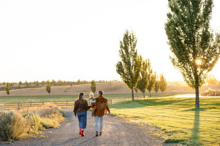 A young family of three walks down the dirt road to the horse stables as the sun sets.