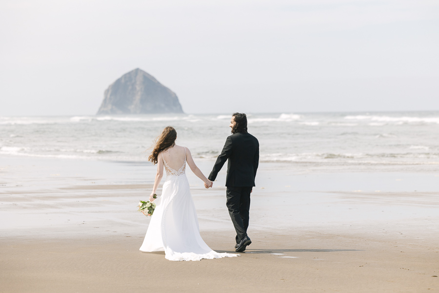 Newlyweds hold hands, smiling as they walk away from the camera down the sandy Oregon beach. 
