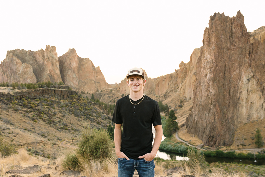 High school senior boy in ball cap stands smiling at camera with Smith Rock in the background.