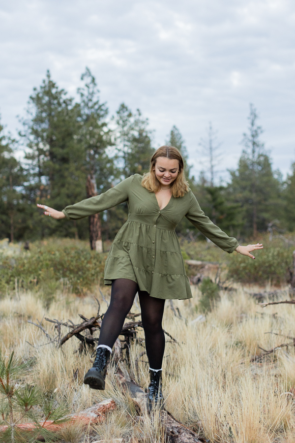 Girl in green dress and black tights balances on downed tree.