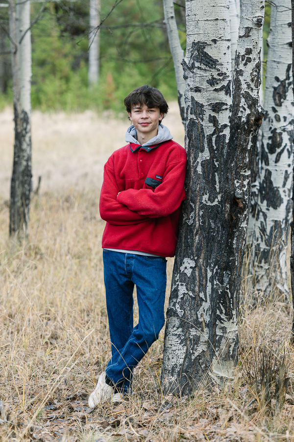 High school senior wear red fleece and denim jeans leans on a tree.
