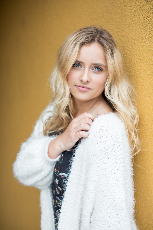 Senior girl with blonde hair leans against yellow colored wall.