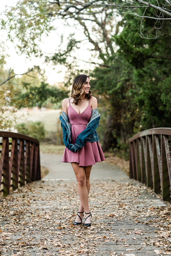 Senior girl in mauve colored dress and denim jacket stands on bridge