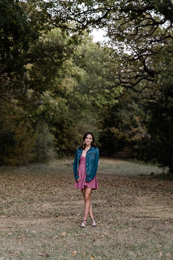 Senior girl in mauve colored dress stands in meadow surrounded by trees.