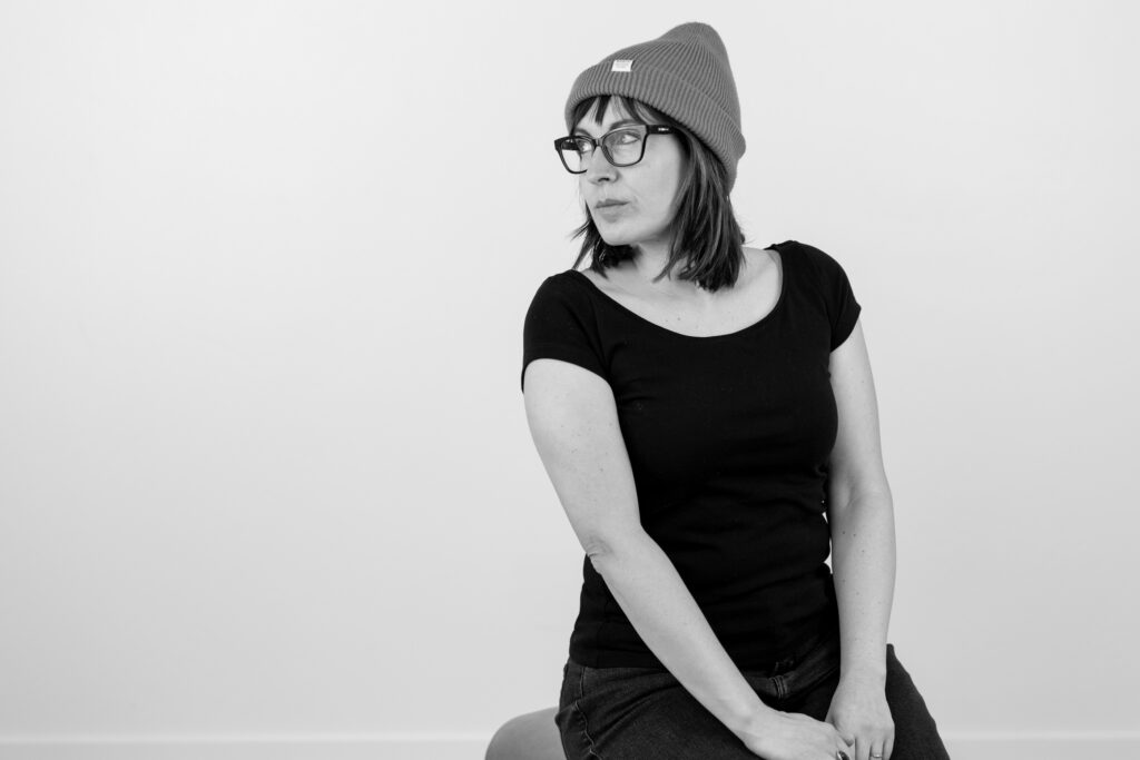 Black and white photo of gwen in a studio. Wearing a beanie and black rimmed glasses.
